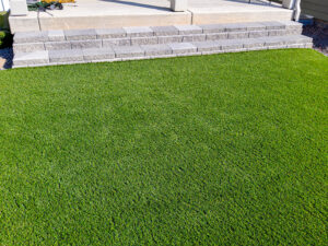 Artificial grass lawn in front of gray stone block steps leading to a concrete patio.