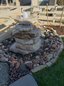 Tiered stone water fountain in a rock-lined circular garden next to grass and fenced yard.